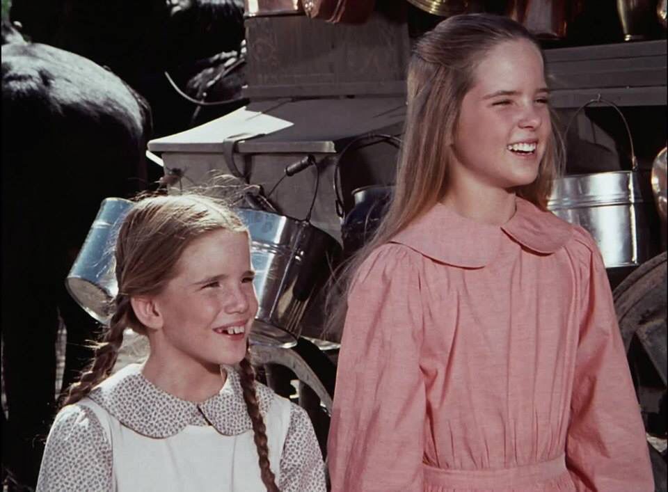 The iconic sibling duo Laura and Mary Ingalls stand smiling in front of a horse-pulled wagon.