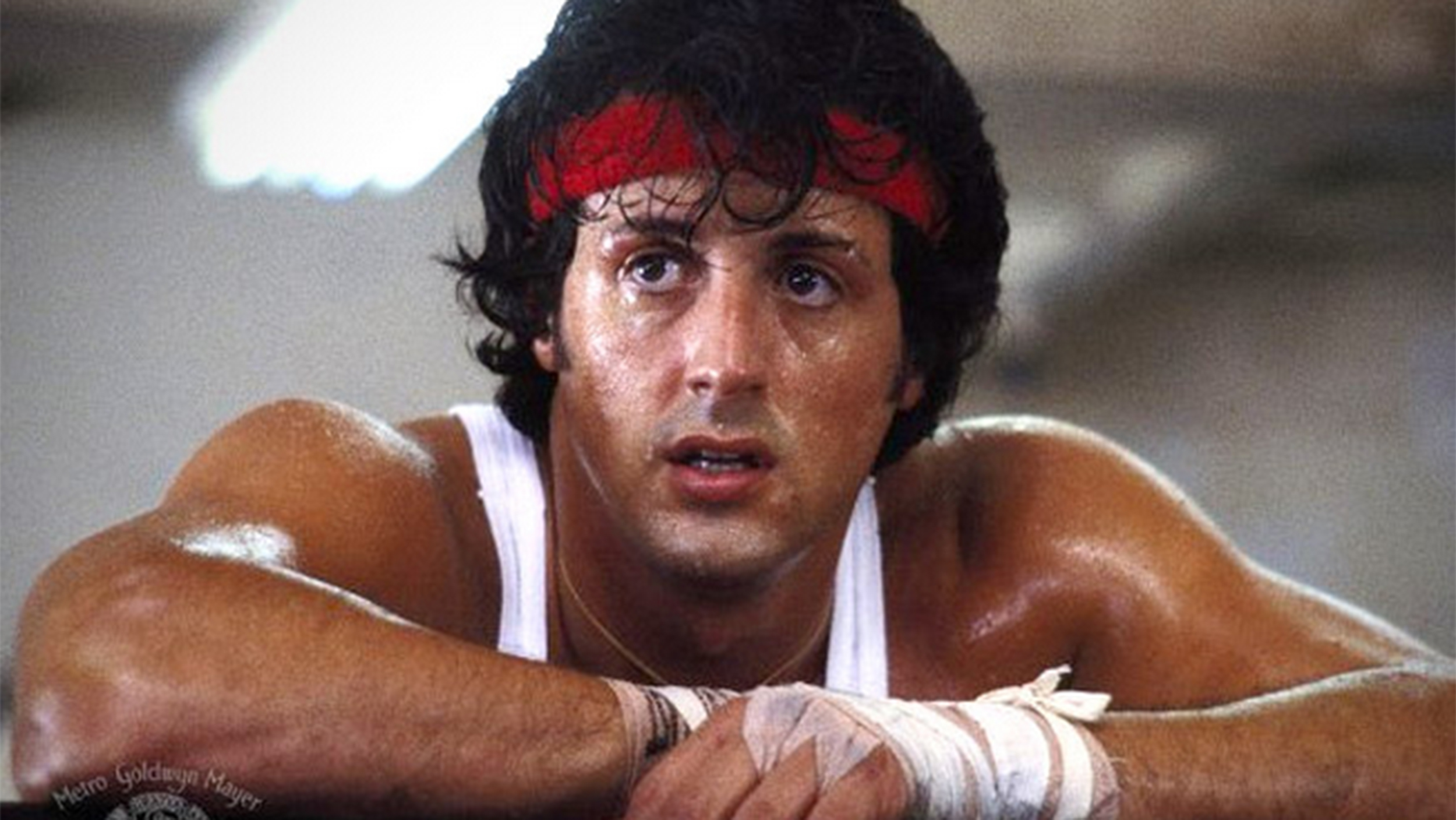 A sweaty Rocky leans forward wearing a red headband. His hands are wrapped in tape for boxing. 