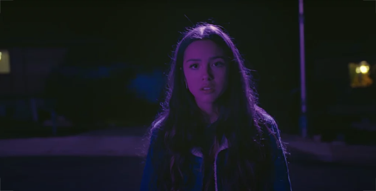 Olivia stands in the center of a street, illuminated in purple light. She stares sadly into the camera.