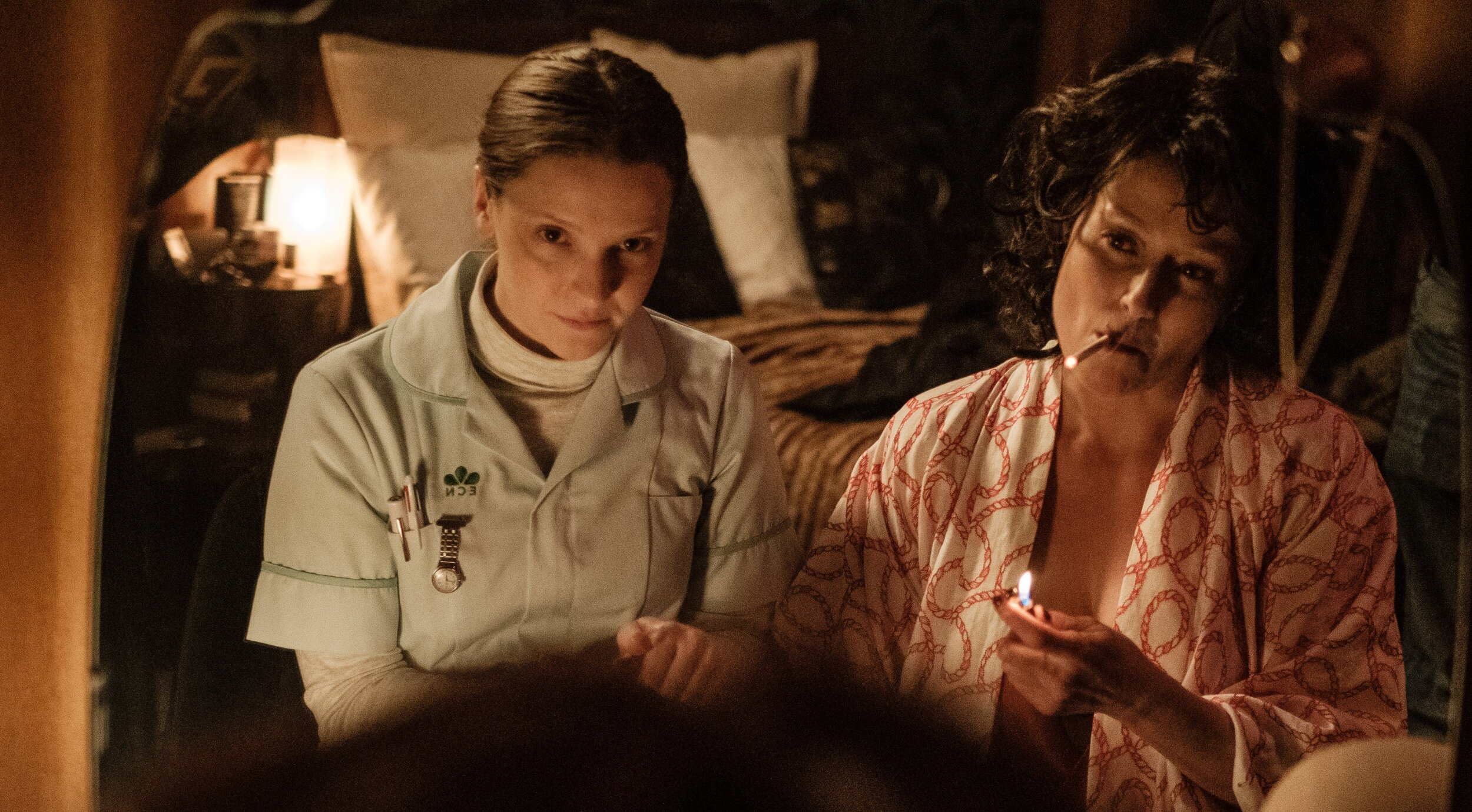 Maud and Amanda sit together at Amanda's vanity mirror. Maud is dressed in her nursing clothes while Amanda is in a lounging robe and smoking a cigarette. 