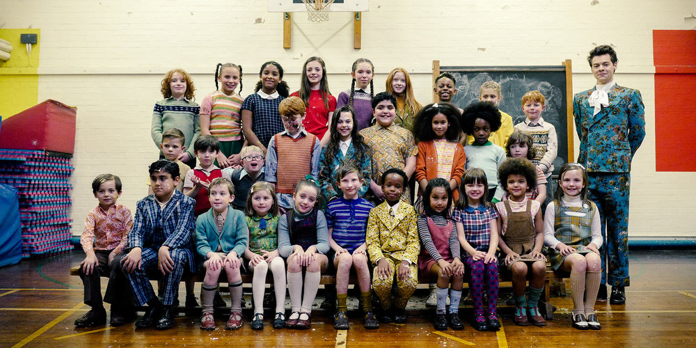 Capture from the "Kiwi" music video of Harry standing beside school children in a school gym.