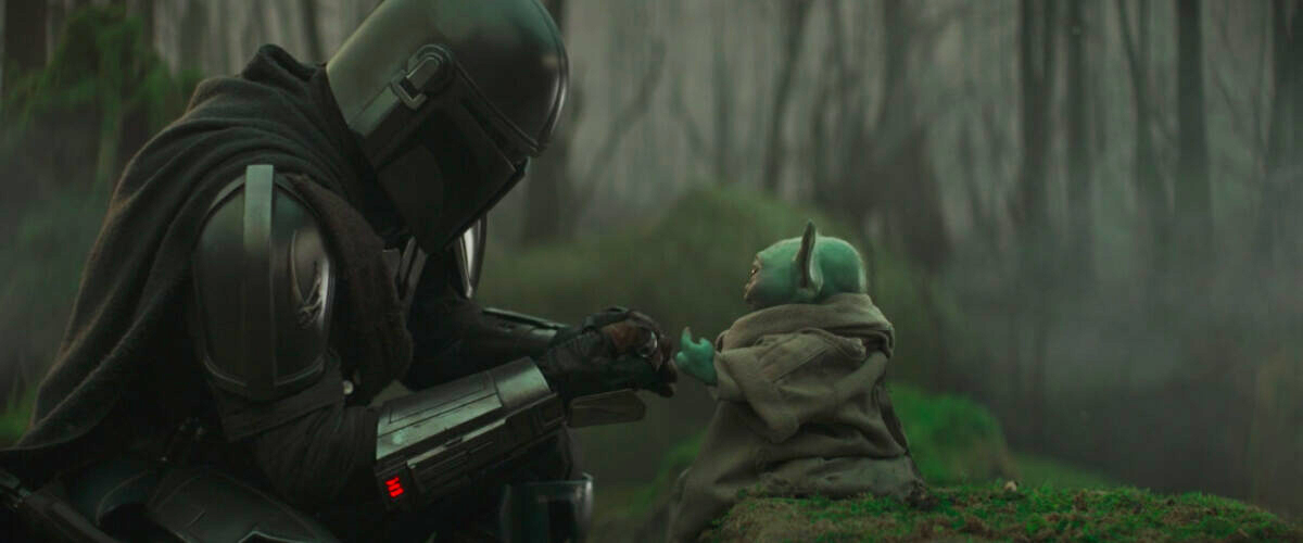In a wooded area, Din Djarin, an armored Mandalorian, kneels in front of Grogu, a large eared alien. They are close to each other and both are reaching hands out. Din is handing Grogu a small metal ball. The two are sharing a familial moment, and it is apparent Din is feeling a sense of pride commonly attributed to fatherhood. 