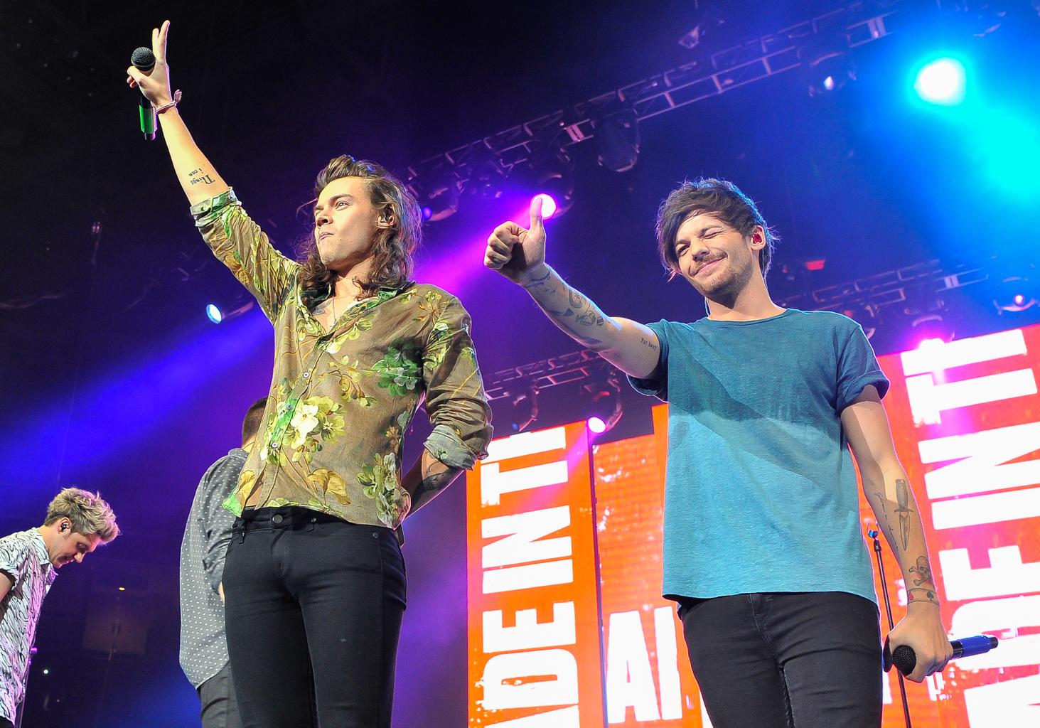 Harry Styles and Louis Tomlinson giving a peace sign and thumbs up to their fans during a concert.