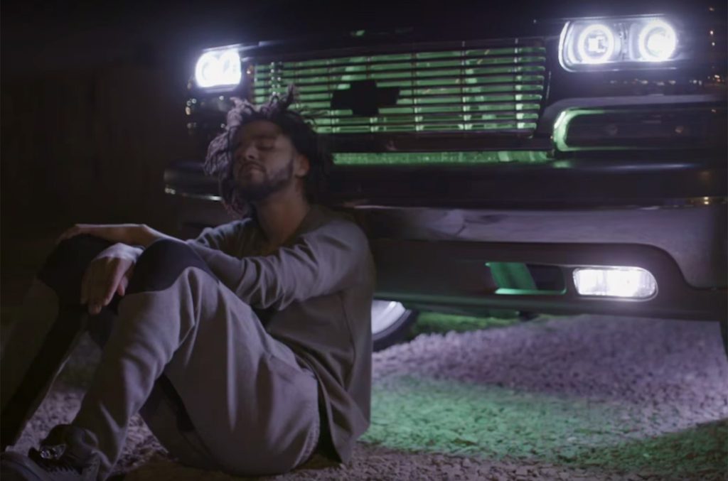 J. Cole sitting in front of a car for the trailer of his documentary.