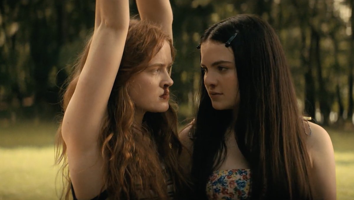 Ziggy Berman (Sadie Sink) with a bloody nose has her arms tied as she hangs from a tree while Sheila (Chiara Aurelia) stands right next to her.