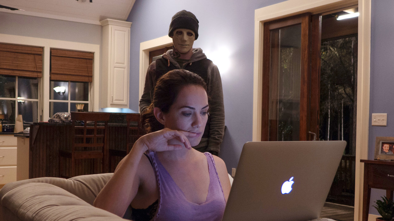 Maddie (Kate Siegel) is on her laptop while The Man (John Gallagher Jr.) is standing behind her.