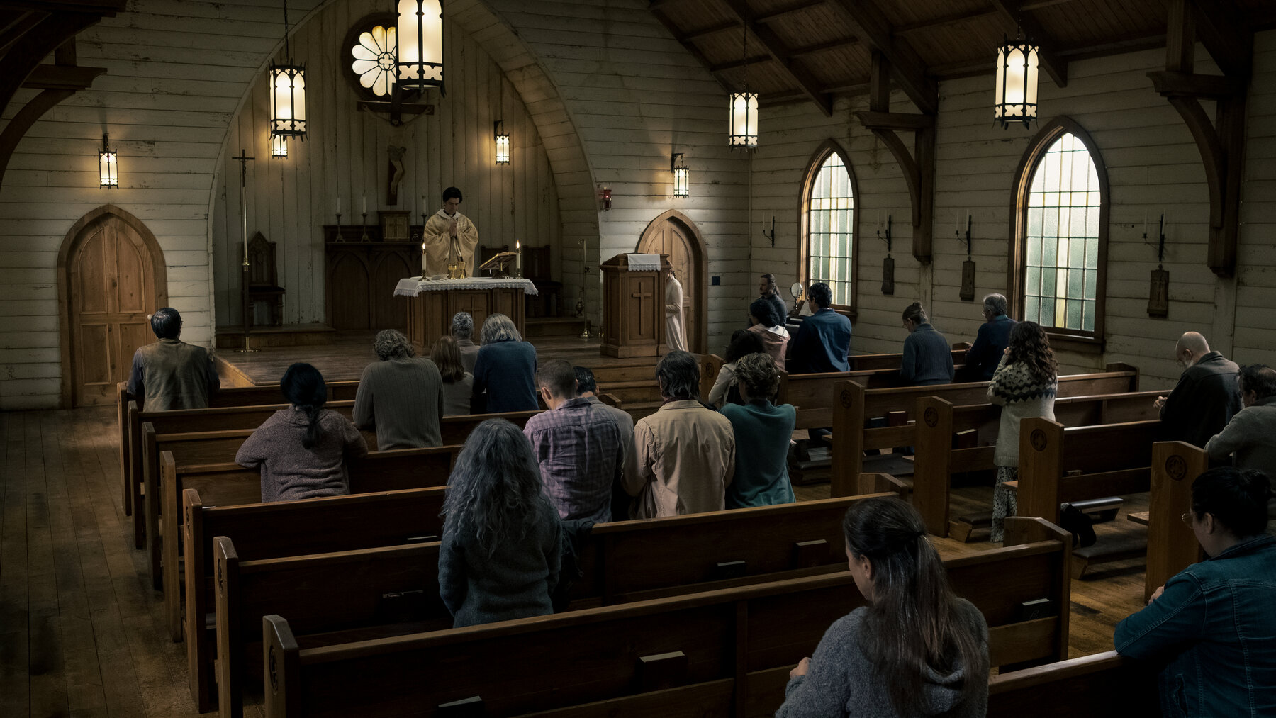 Father Paul stands in front of the congregation in a wide shot of the church. Midnight Mass. Season 1, Episode 1: "Book I: Genesis". 2021. Netflix Entertainment. 