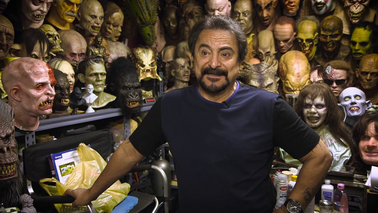 Tom Savini stands in a room filled with character designs of all of the horror villains he has worked on or helped to work on throughout his career.