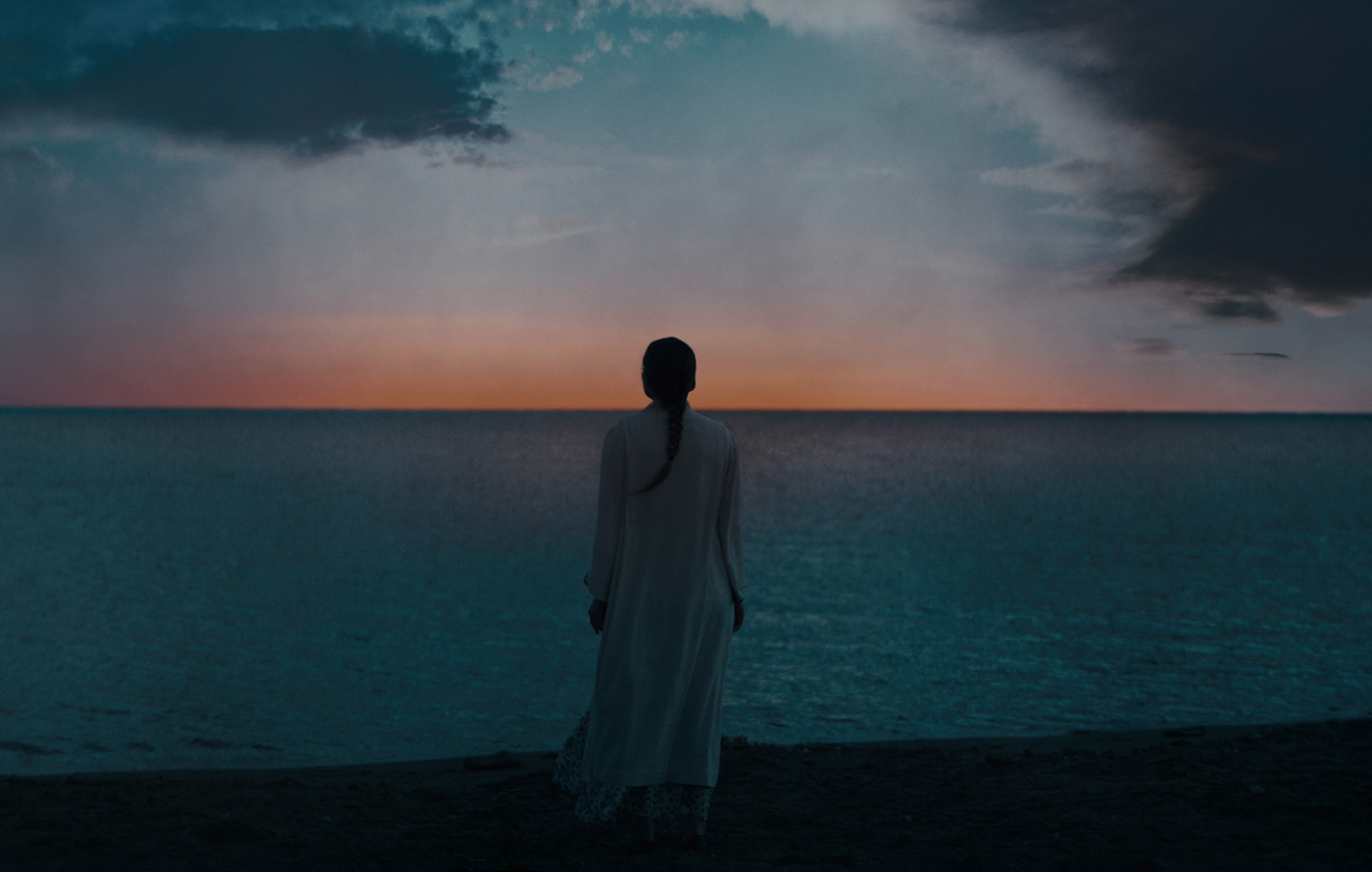 Bev Keane stands in center frame on the shore of Crockett, looking out at the very beginnings of a sunrise. Midnight Mass. Season 1, Episode 7: "Book VII: Revelation". 2021. Netflix Entertainment.