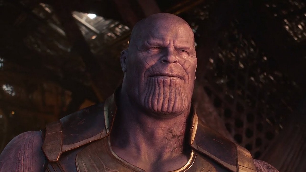 Villains: Thanos, a purple alien, stands in a ruined hut. We can only see him from the shoulder up. he is scarred on his left side. On of the MCU's Villains.