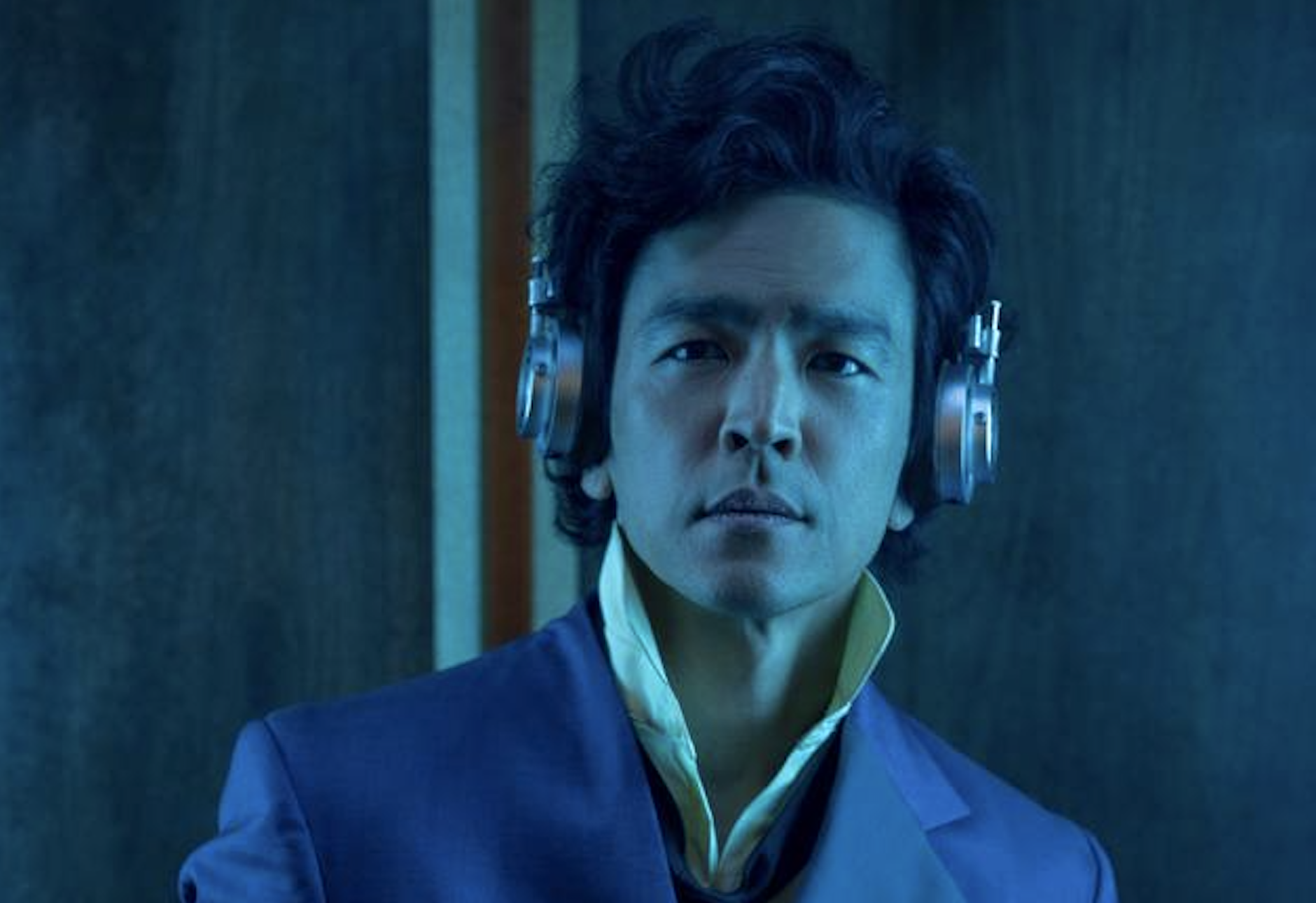 John Cho as Spike Spiegel in the 2021 Netflix Cowboy Bebop adaptation. He's wearing Spike's iconic blue suit with a yellow collared shirt. He also has a pair of grey headphones on. The picture has a blue filter.