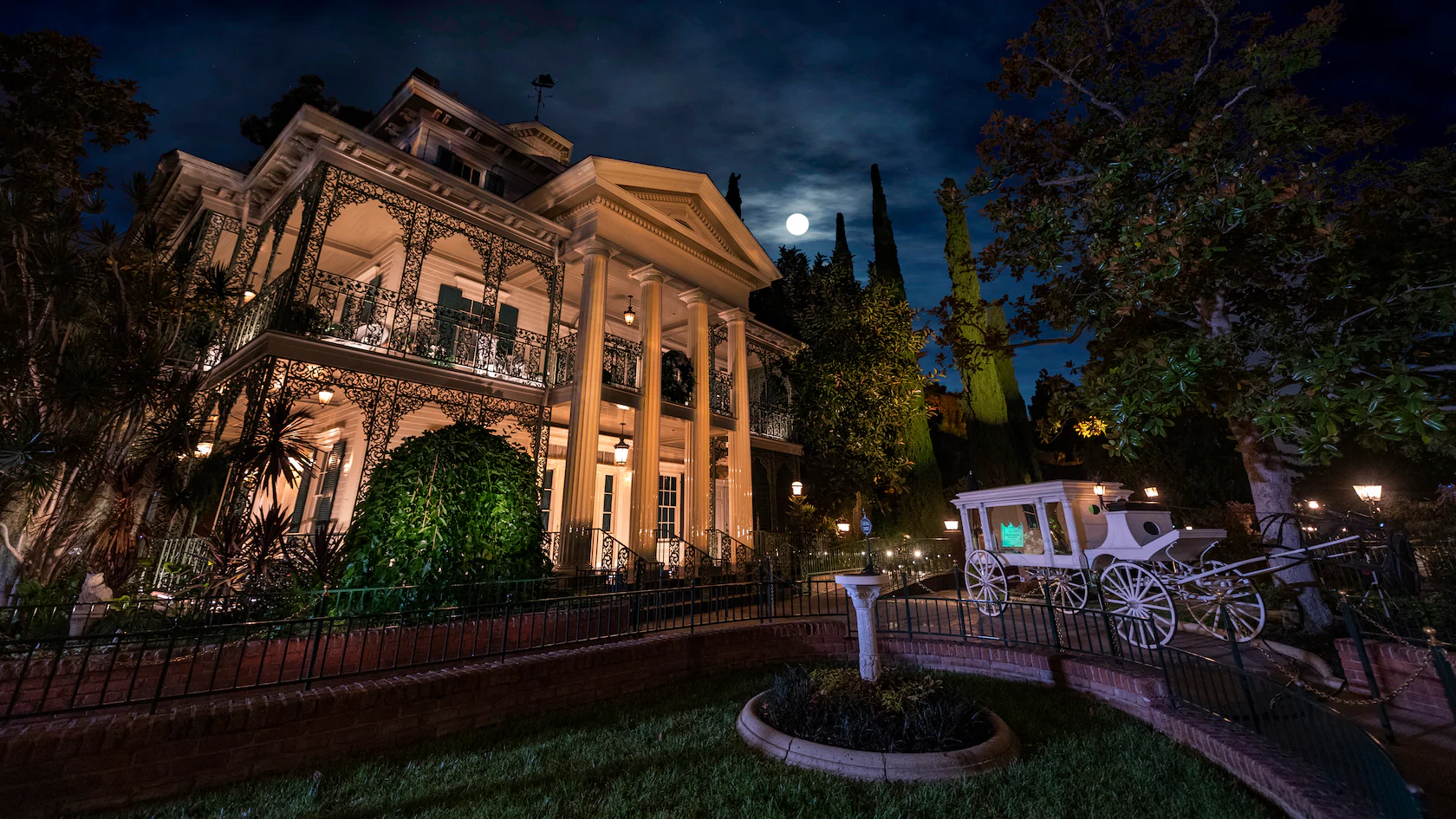 The Disneyland version of The Haunted Mansion looms in light of a full moon. The mansion is fully illuminated in the darkness, giving it a ghostly aura. 