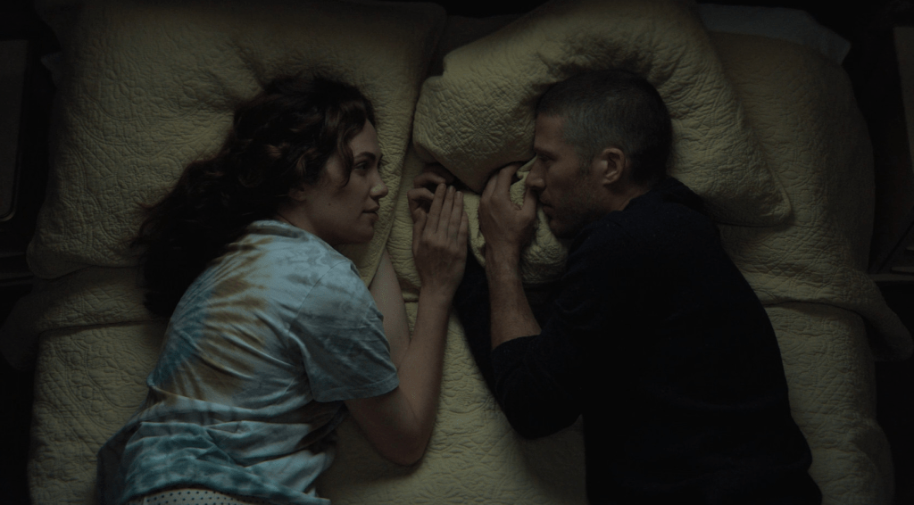 Erin Greene and Riley Flynn lay facing each other on a bed from an overhead shot. Midnight Mass. Season 1, Episode 4: "Book IV: Lamentations". 2021. Netflix Entertainment. 