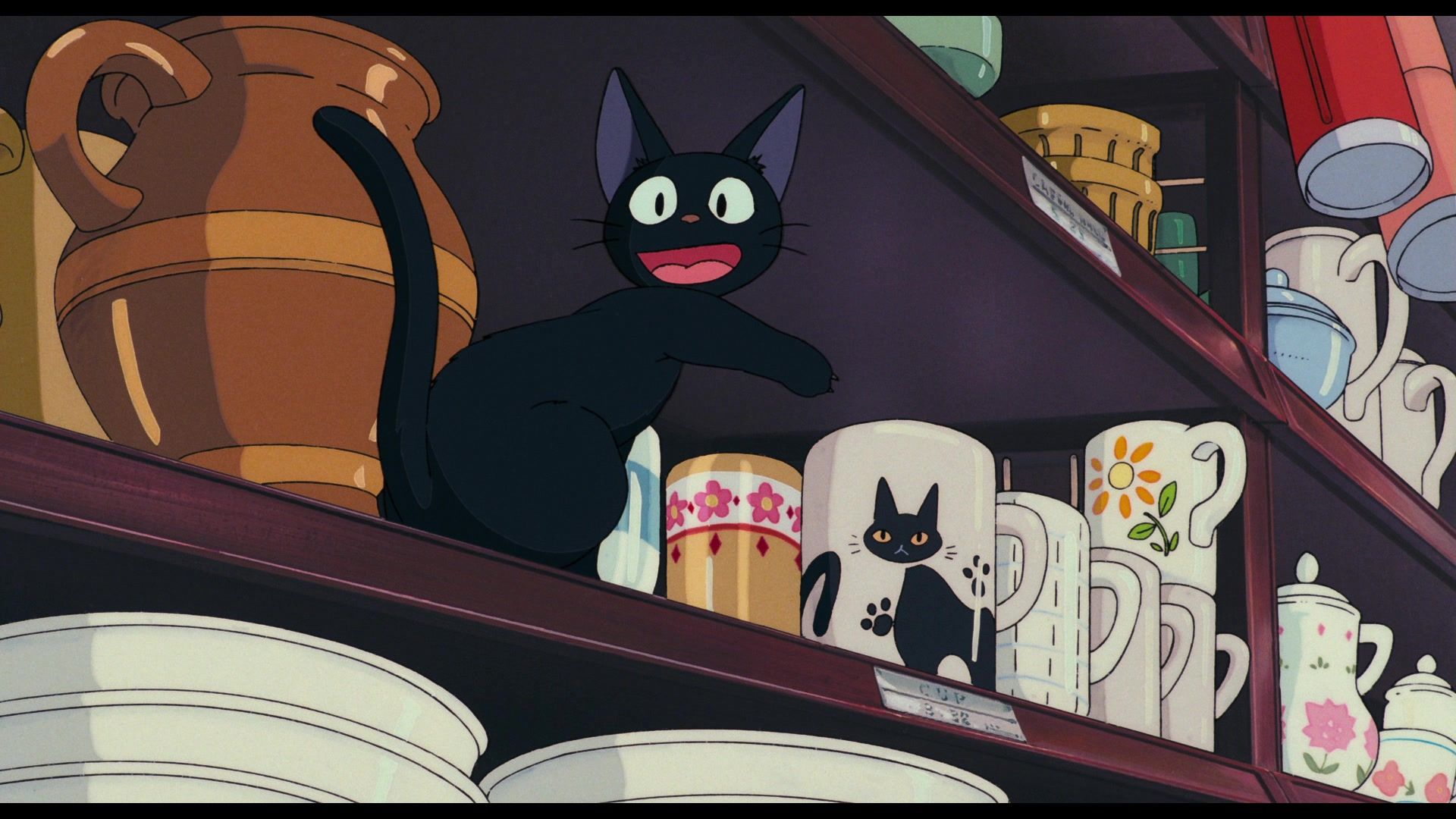 Jiji sits on a shelf filled with various mugs and vases. He points happily at a mug that shows a black cat that looks like him. 