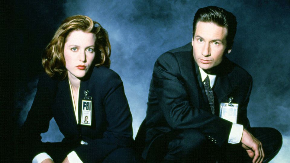 Mulder and Scully sit side by side in a promotional image for The X-Files.