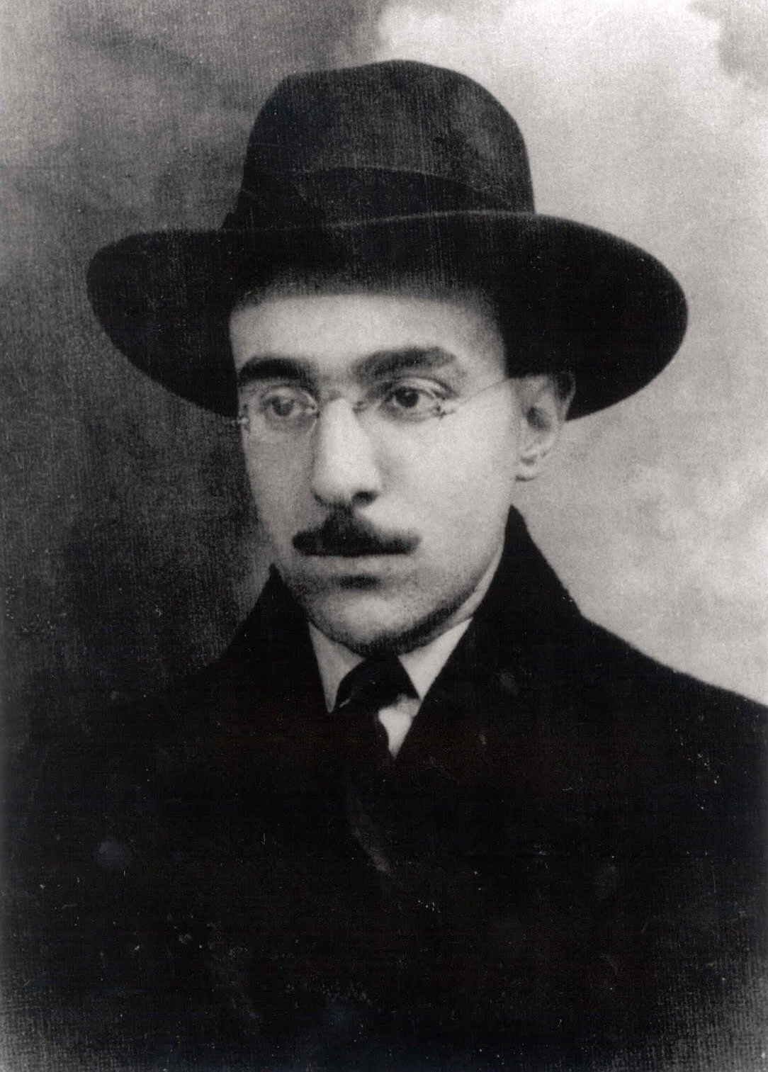 A black and white photo of Fernando Pessoa as a grown man, though still fairly young. He is in a suit and tie, wearing a full-brim hat, and glasses. He has a mustache that covers his upper lip, and thick eyebrows.