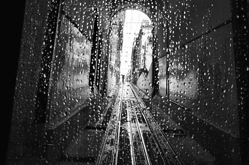 A black and white photo of a road in Lisbon. Streaks of rain mark the photo, which depicts trolley tracks in the foreground and a person holding an umbrella in the background. 