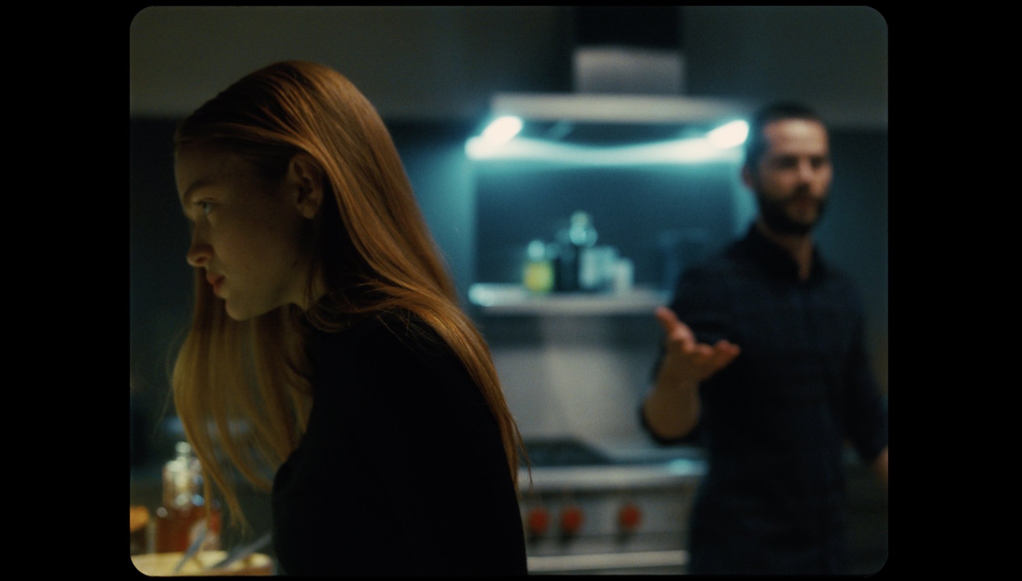 In a medium shot, Sink stands in the foreground colored in yellow lighting with an irritated look on her face. O'Brien is out of focus, a few steps behind her, with an outstreched hand colored in artifical blue lighting. 