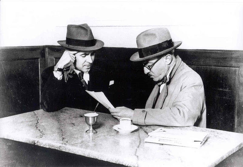 A black and white photo of Pessoa with colleague, Costa Brochado, at a cafe in Lisbon. They are both wearing brimmed hats, suits, and ties. They are looking down at a piece of paper. There is a cup of coffee and sugar bowl on the table. 