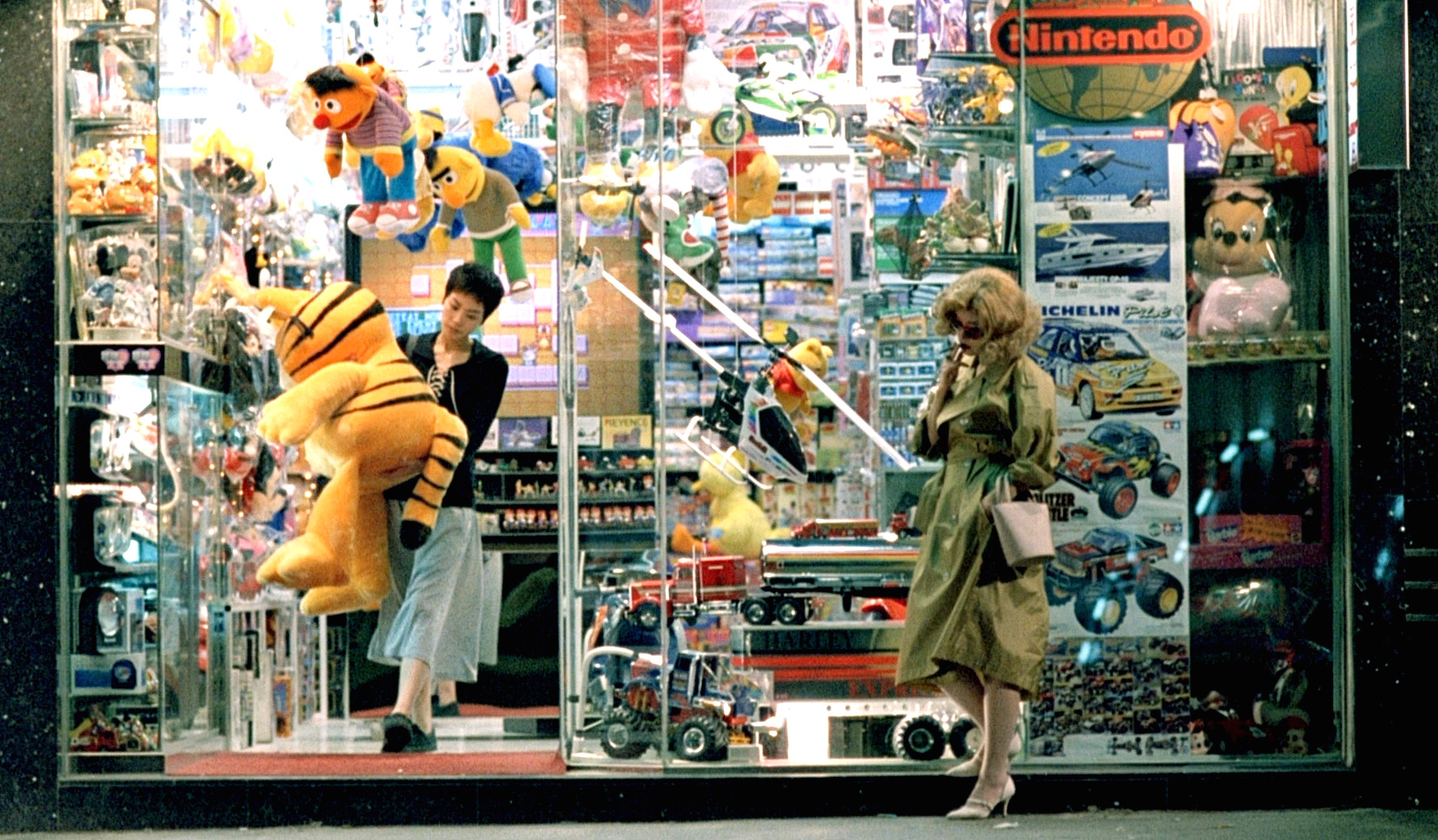 This is a film still from Chungking Express. A woman in sunglasses and a blond wig stands outside a toy store in Hong Kong smoking a cigarette. Another woman is walking out of the store with a large stuffed animal of Garfield. The store is packed with lots of 90s era toys.