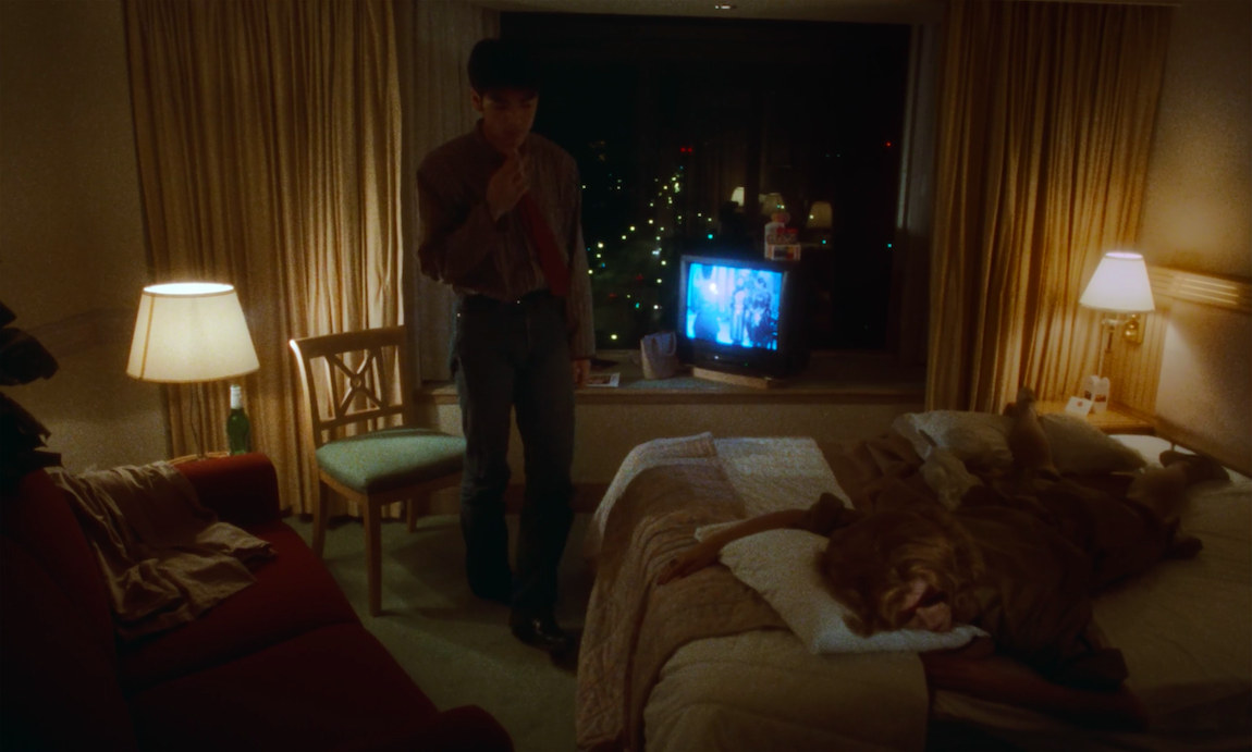 He Qiwu stands in a hotel room with the television on while the blond woman sleeps in bed. The Hong Kong skyline is shown in the window. It's nighttime. 