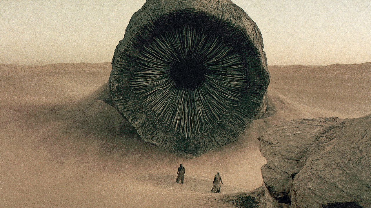 An image of a worm from the 2021 'Dune' rising from the desert in front of two dwarfed figures.