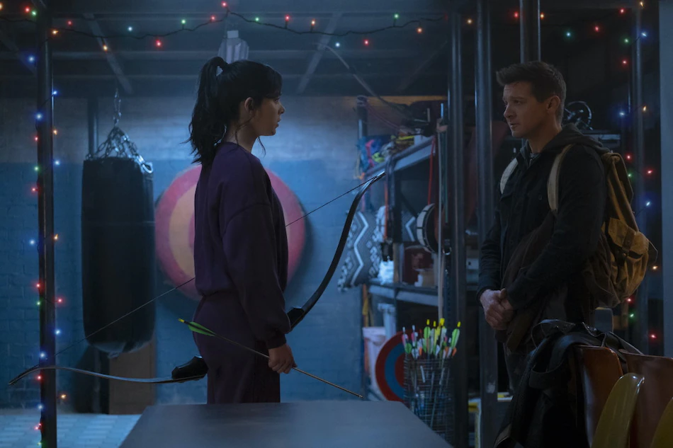 Kate Bishop and Clint Barton stand in Kate's apartment. It is decorated for Christmas. Kate holds a bow and Clint is wearing a backpack.