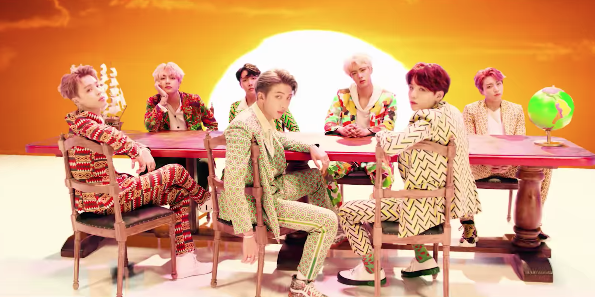 BTS sit in colorful suits around a table. 