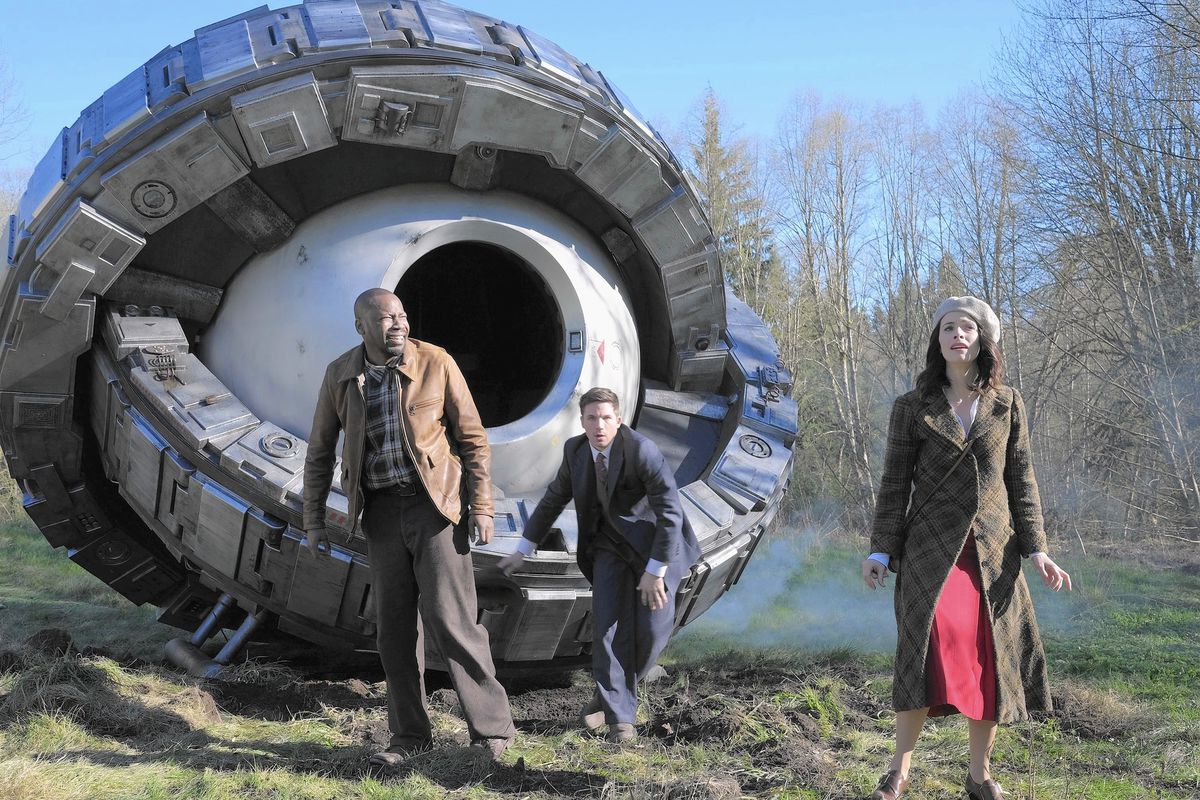 Three people are in the middle of the woods, looking disoriented. There is a large time machine behind them, which looks like an orb with two mechanical bands that rotate around it.