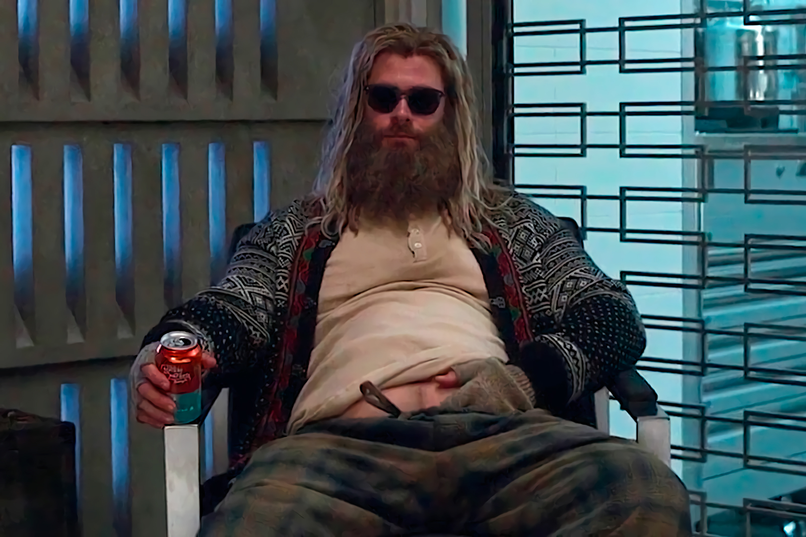 Thor sits back in a chair with a hand on his exposed belly, and a soda or alcoholic beverage in his other hand. He wears sunglasses indoors and his clothes look unwashed. 