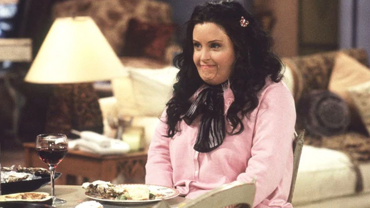 "Fat" Monica smiles and sits at a dinner table with a glass of wine and a plate of food in front of her. 