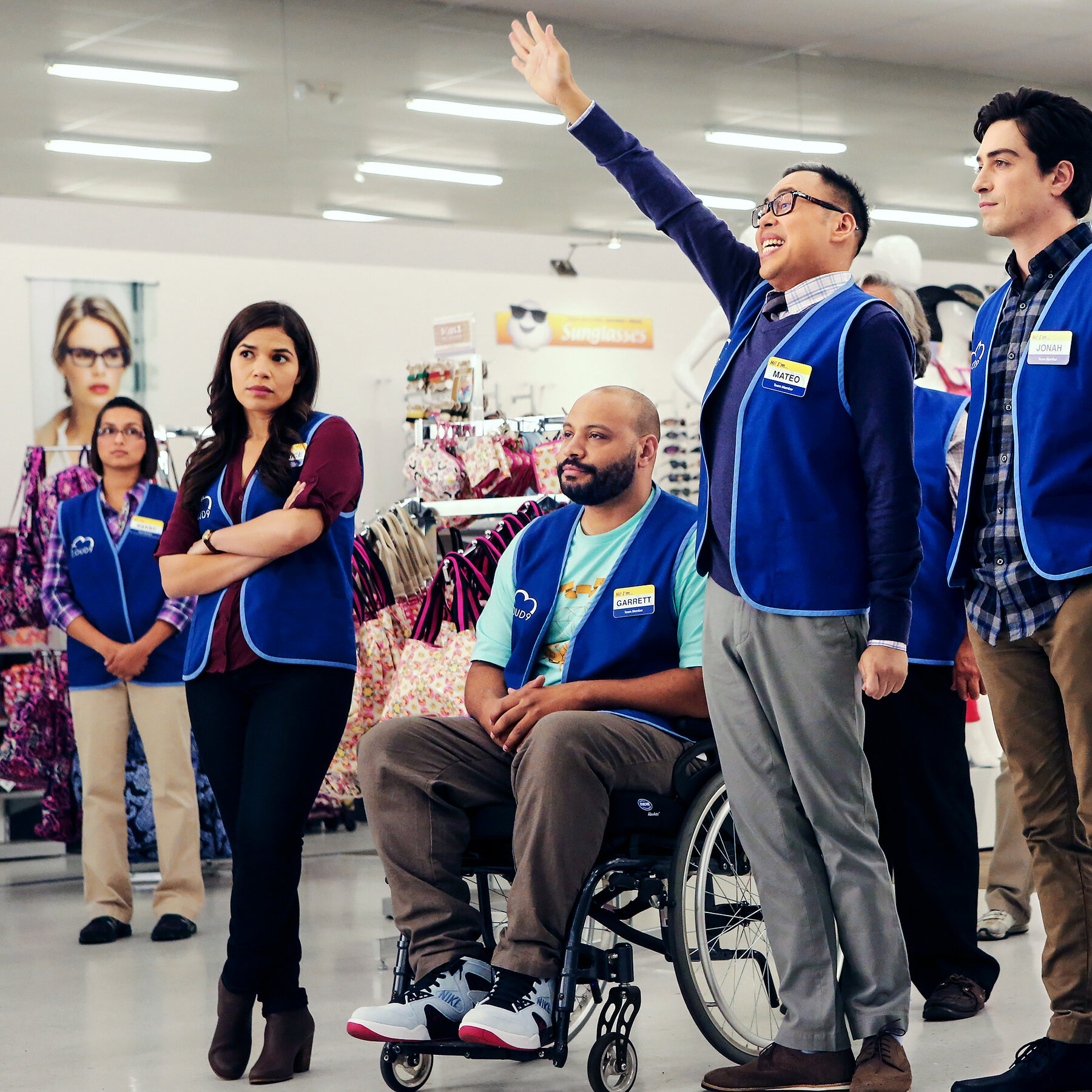 Amy (America Ferrera), Garrett (Colton Dunn), Mateo (Nico Santos), and Jonah (Ben Feldman) watch something offscreen. Amy is standing with her arms crossed, Garrett sits in his wheelchair with his hands in his lap, Mateo is standing with a hand raised in the air and Jonah stands with his hands in his pockets. Superstore. 2015-2021. NBC.