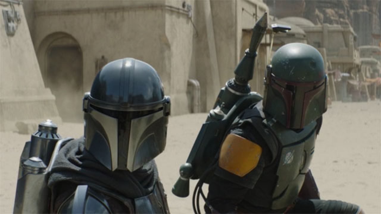 The characters The Mandalorian and Boba Fett kneel down avoiding blaster fire on the streets of Tatooine.