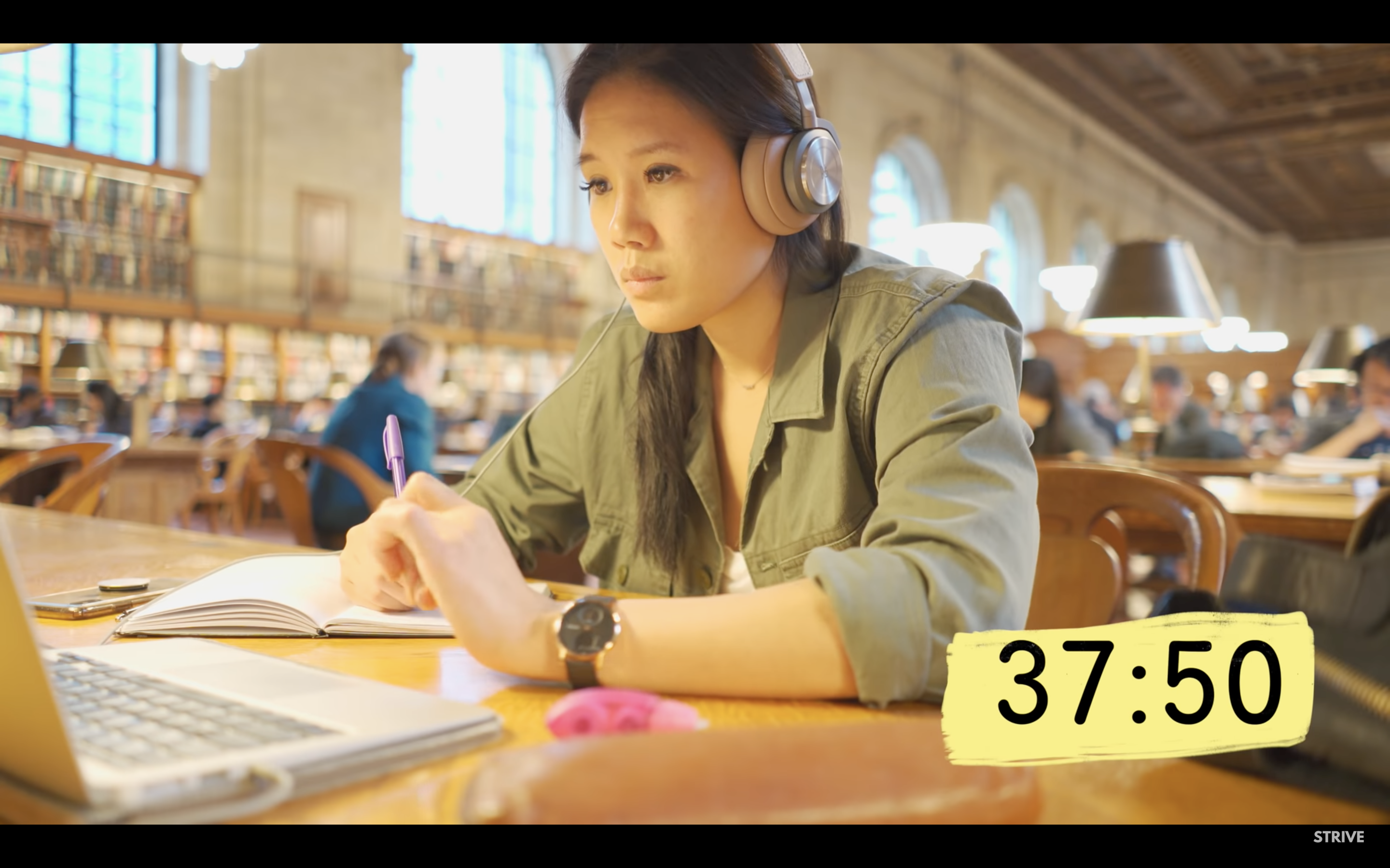 Screenshot of a library "study with me" video posted by TheStrive Studies. A woman is sitting at a table in a library while taking notes and looking at a computer, and a timer is in the bottom right corner of the screen reading "37:50".