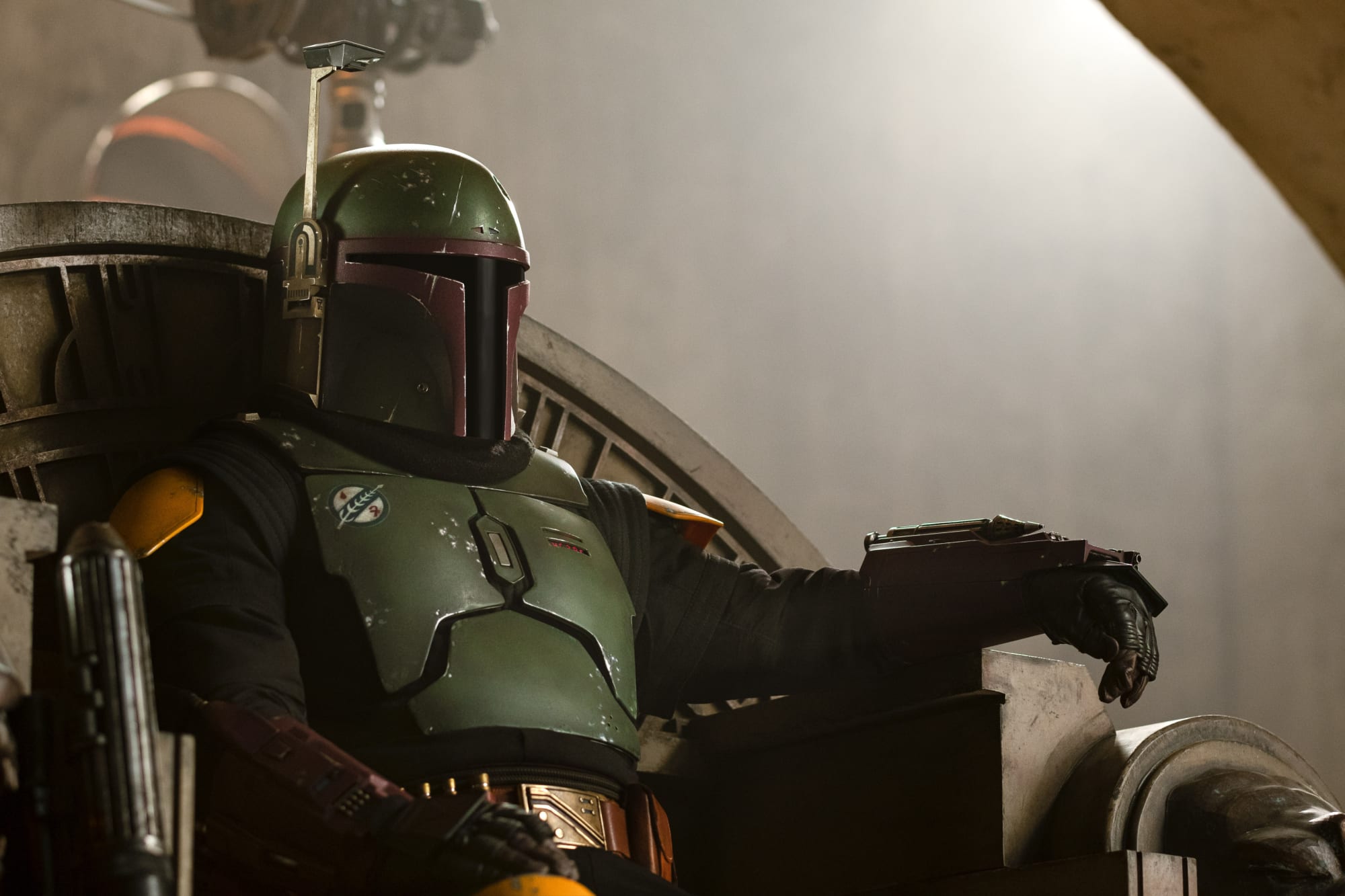 Boba Fett sits on the throne in his palace. He is wearing green armor on a stone throne with a blaster leaning on one of the arms.