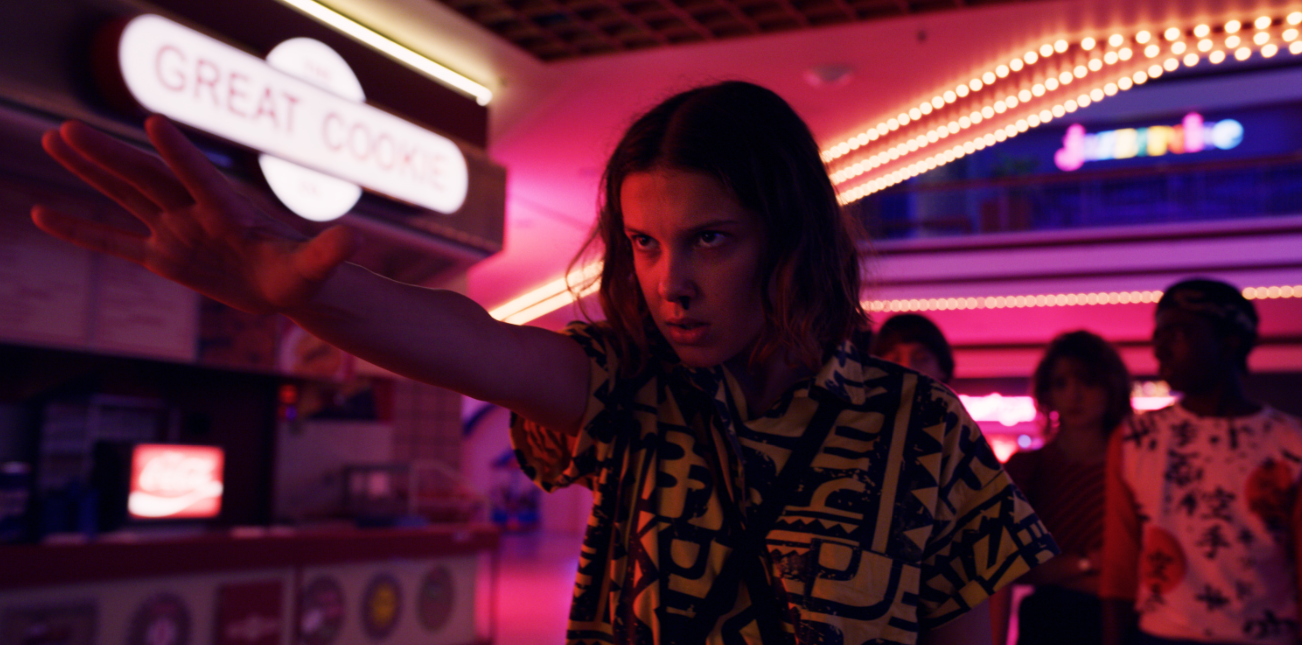 Stranger Things protagonist, Eleven, stands in the neon lighting of the Starcourt Mall with her hand outstretched as she concentrates on her powers. 