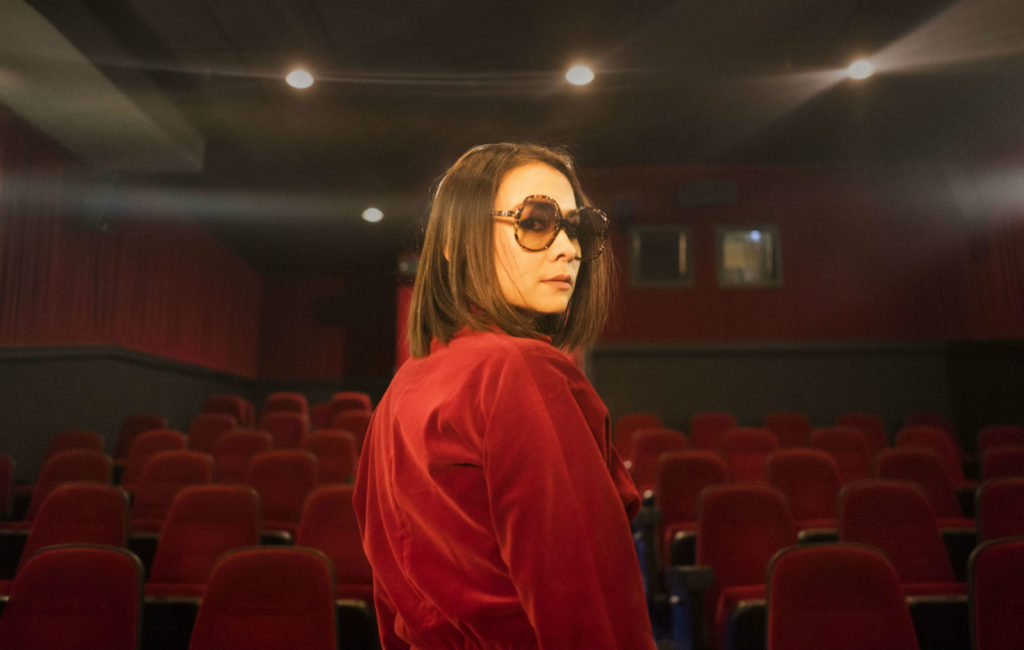 Mitski looks over her shoulder, the backdrop an empty movie theater. (NME. Aug. 2018).