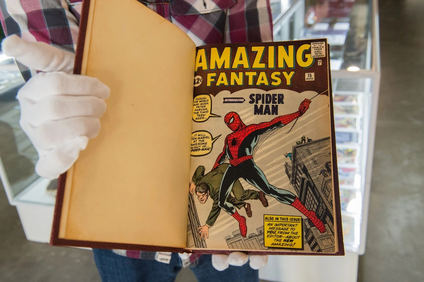 A white-gloved hand gently holds open a protective book that contains a copy of the 1962 Marvel comic wherein Spider-Man makes his debut. The cover reads, "Amazing Fantasy" in bold yellow letters. Spider-Man swings across the center, a string of web in one hand and a flailing man in the other.