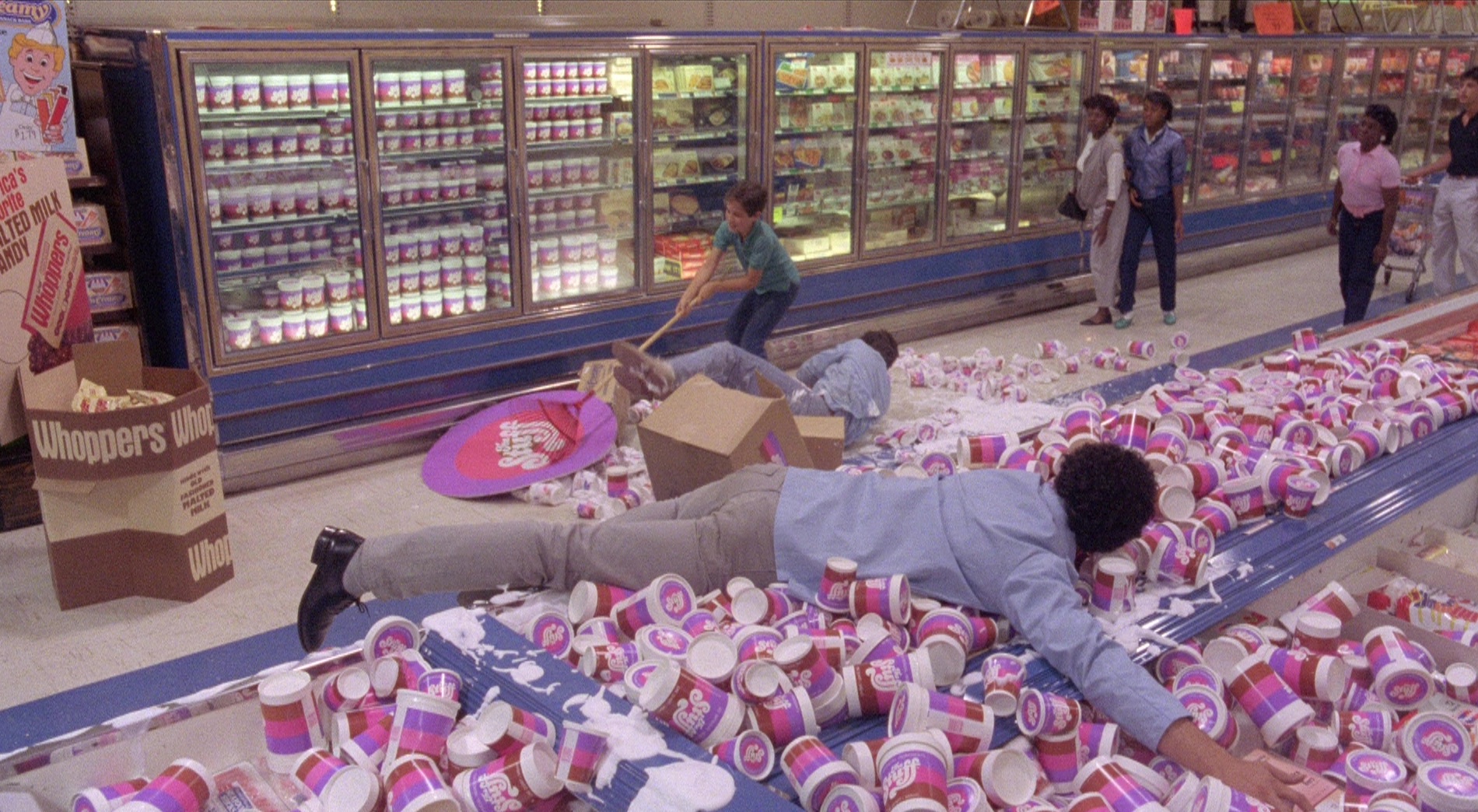 A still from The Stuff that shows the local grocery store overrun with the newest product - The Stuff. Tubs of the ice cream overtake the store as costumers jump and fall over to reach the product.