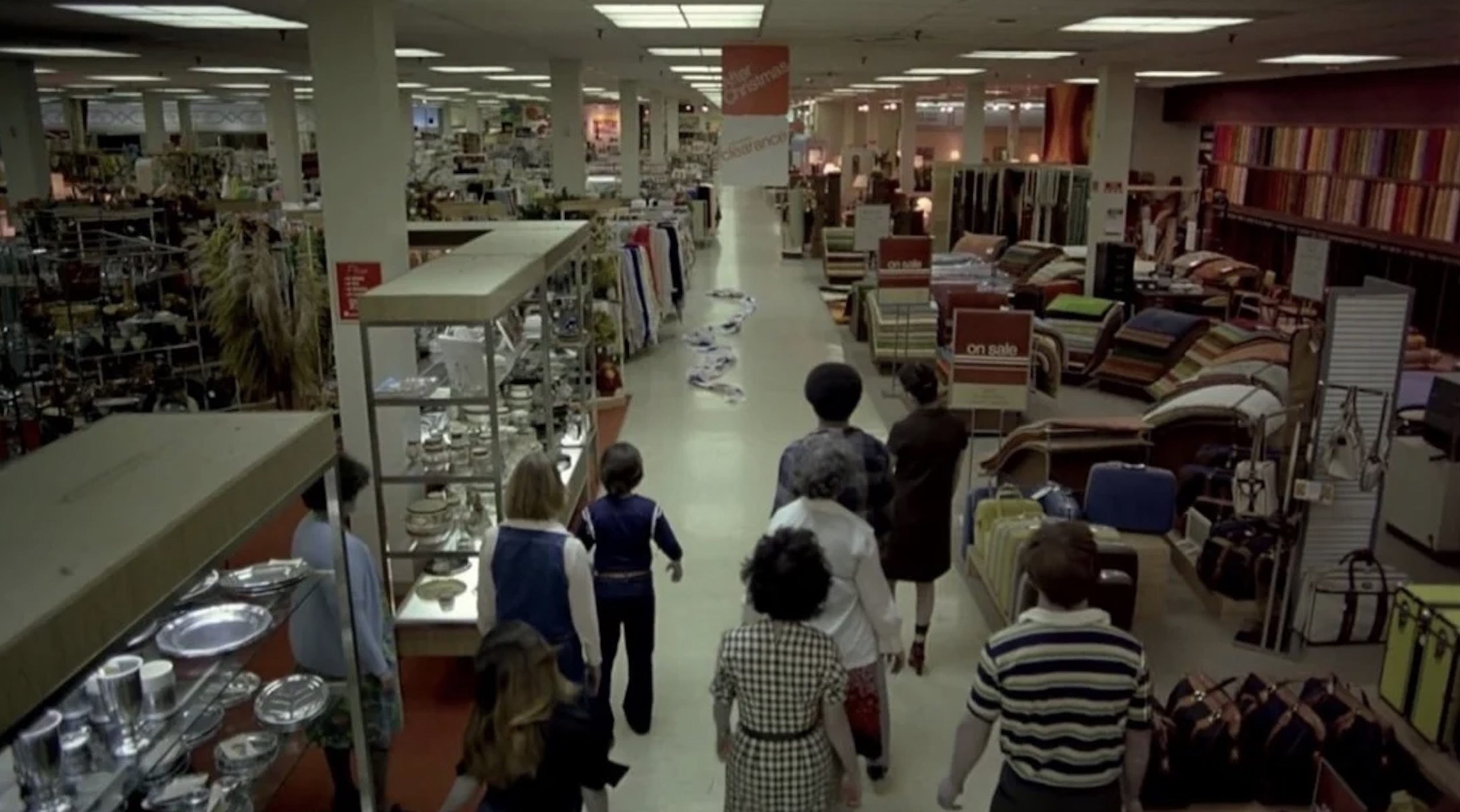 In this frame from Romero's Dawn of the Dead, a group of zombies stand with their backs to the camera in the foreground of the image. The rest of the shot shows an empty department store with a blood-stained cloth lying in the center of the aisle. 