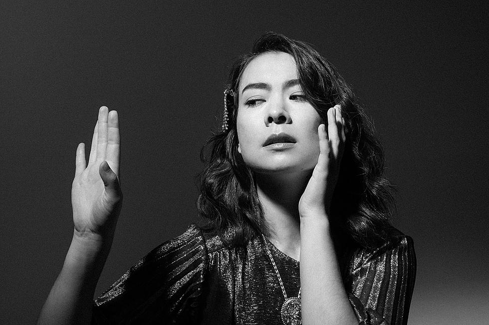 Mitski gazes at her raised left hand, while her right caresses her face for a photoshoot. Image is in black and white. (Yildiz, Ebru. BrooklynVegan. Jan. 2022).