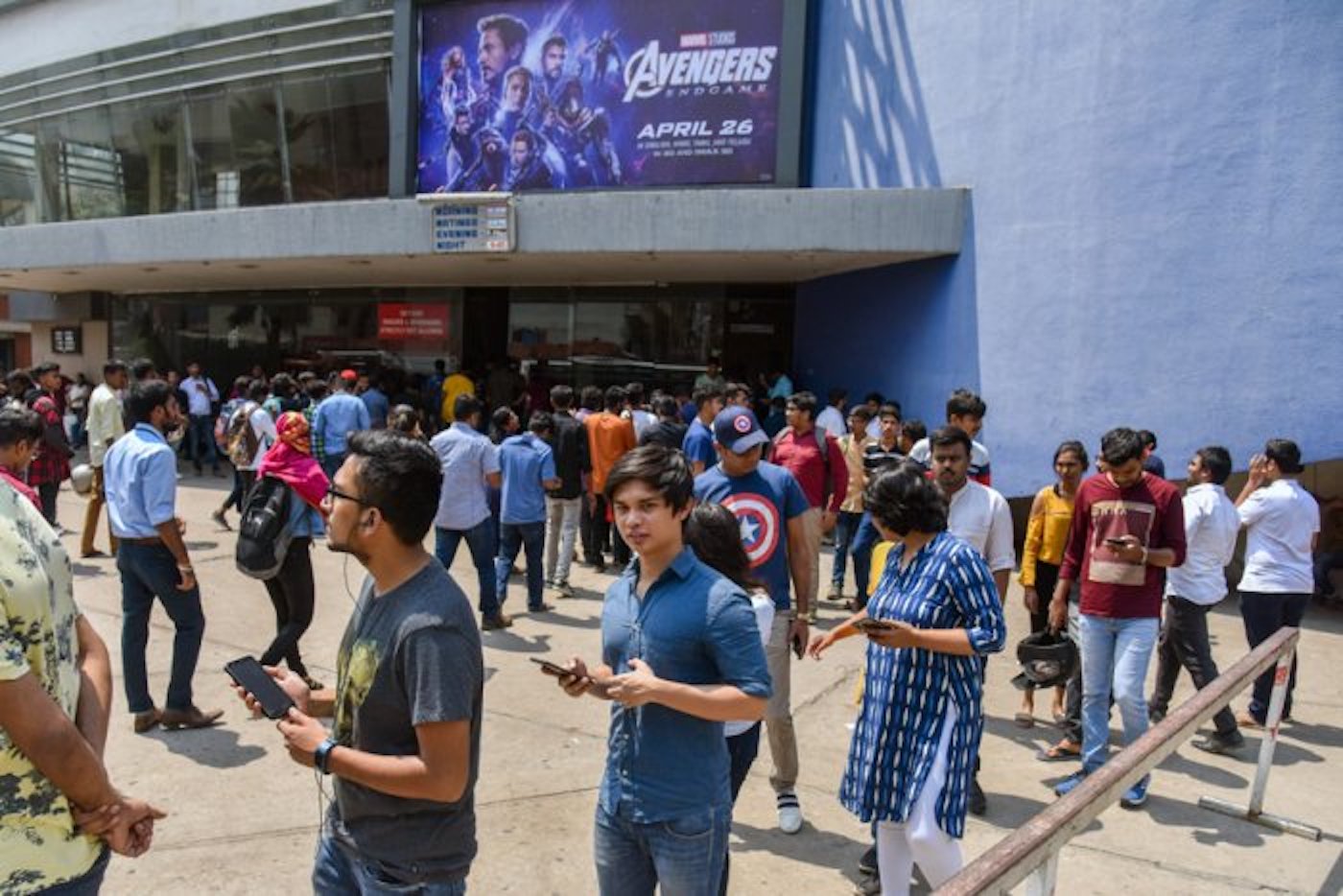 A line of people queues outside of a large blue theater before a showing of "Avengers: Endgame" (2019). 