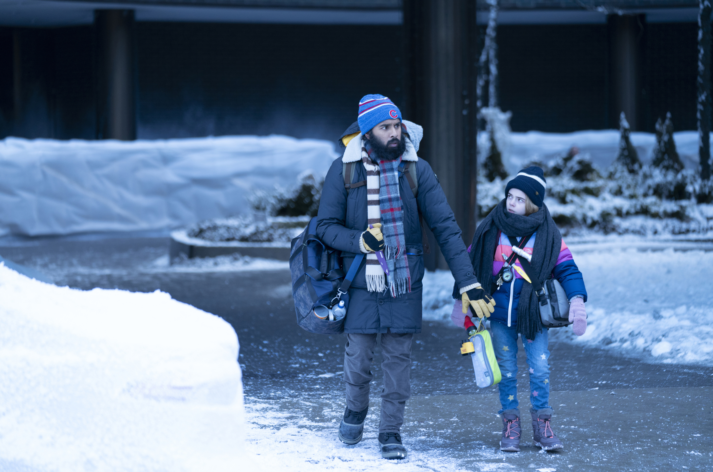 Jeevan (Himesh Patel) and Young Kirsten (Matilda Lawler) walk on a street in the snow, bundled up in many layers.