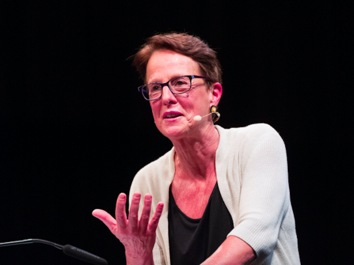 Wendy Brown stands at a podium against a black background as harsh stage lighting shines down on her. She is mid-sentence, one hand gesturing in an upward palm motion. Her brown hair is cut close to her head and she is wearing black rectangle glasses. A mic has been attached over her ear and protrudes to the side of her mouth.