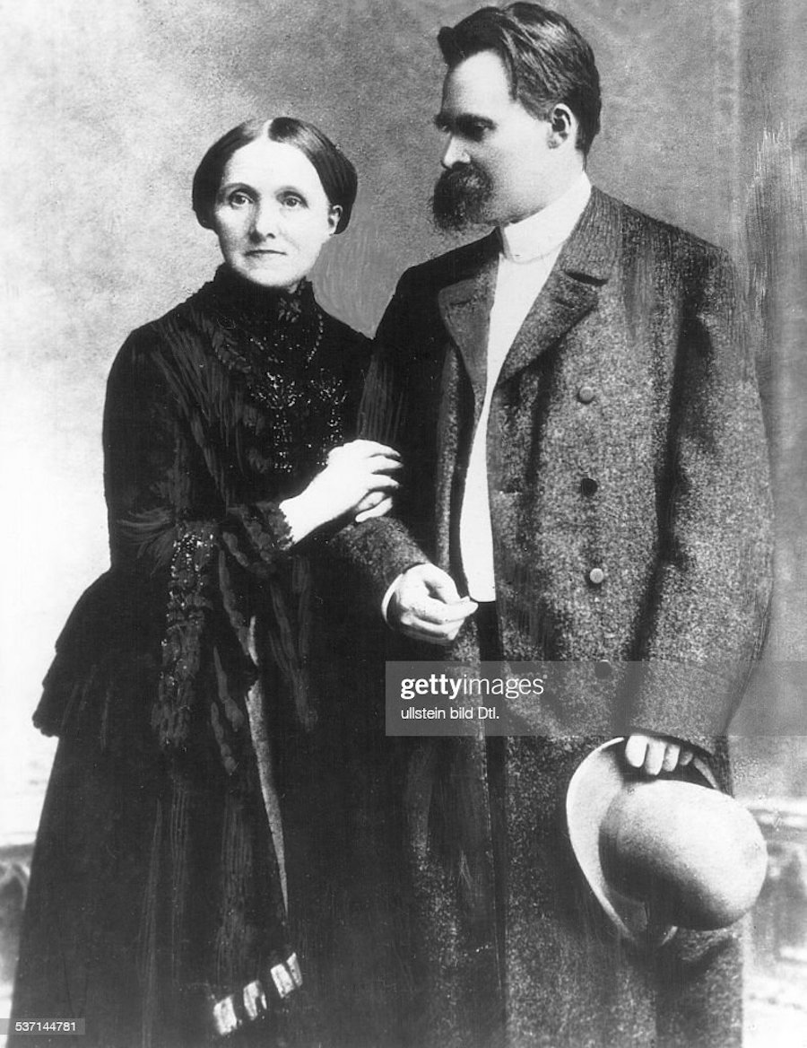 A black and white photograph depicts Fredrich Nietzsche and his mother,  Franziska Nietzsche. Fredrich stands tall, looking down and to the side at his mother who holds his arm. She stairs ahead, past the camera. Both are dressed in formal clothes. 