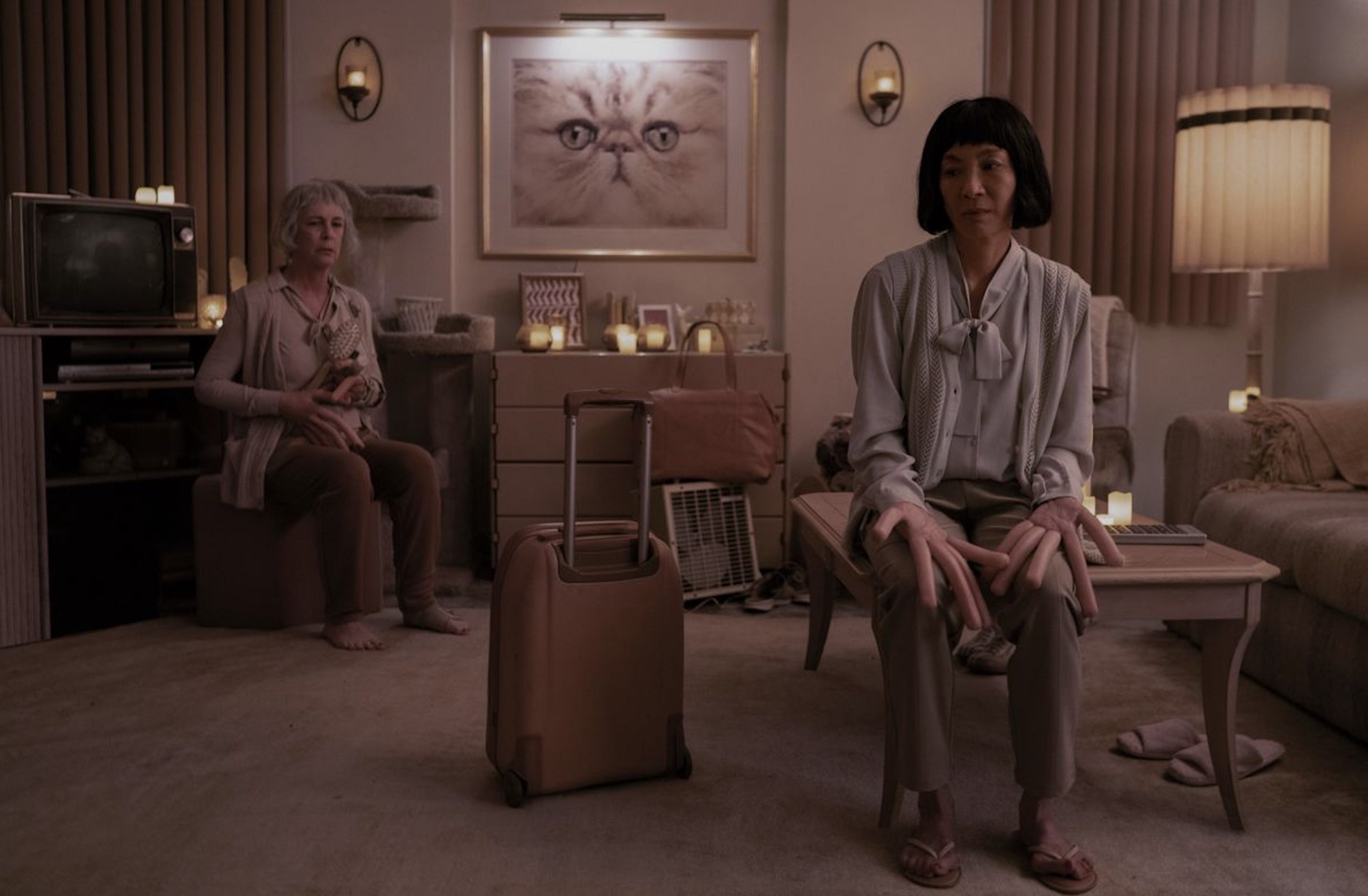 In a clean, peach apartment with a large cat photo framed in the center of the image, Evelyn sits on a coffee table with her hot dog finger hands resting on her knees. Jamie Lee Curtis's character sits in the background looking apprehensive.