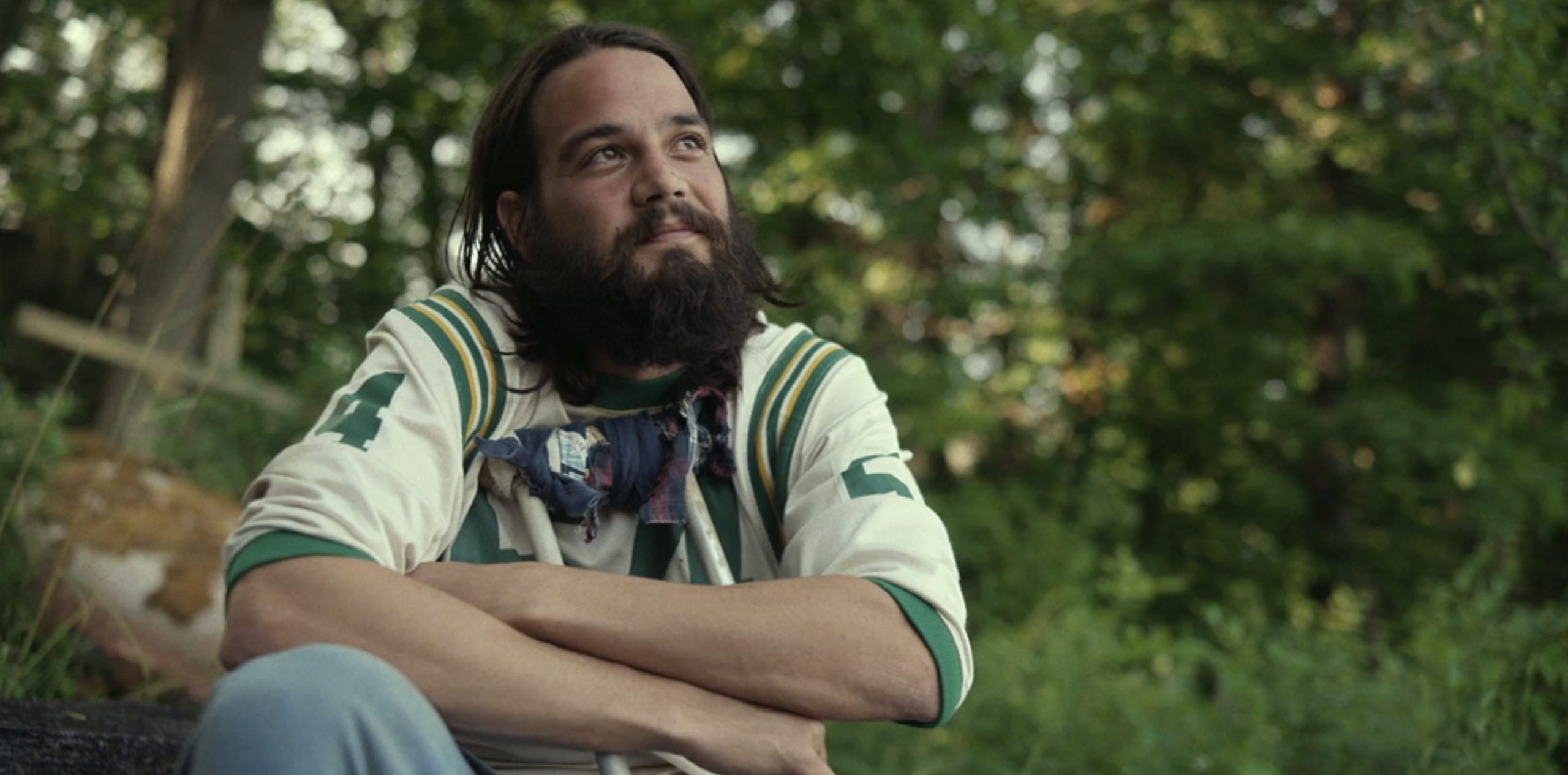 Tyler (Daniel Zovatto), a bearded man with long hair wearing a sports jersey, sits outside. 