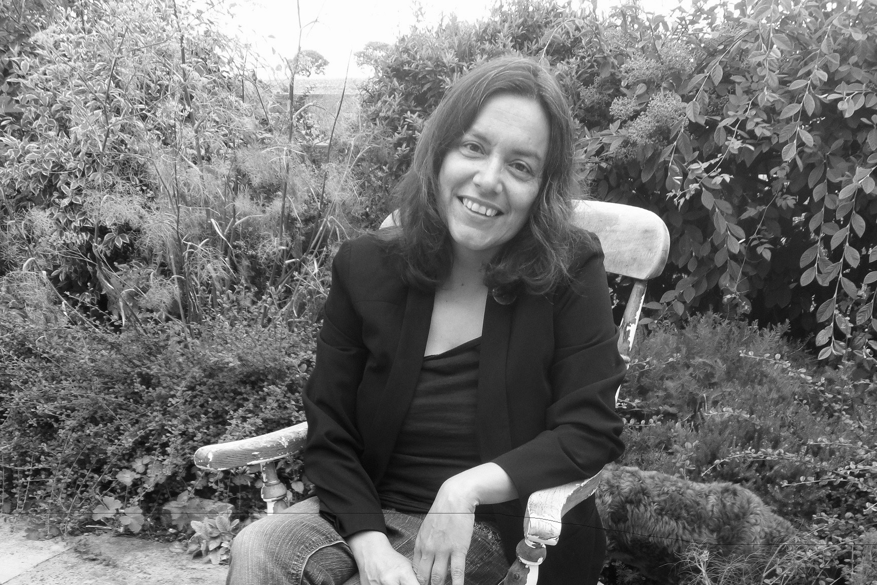 A black and white photograph shows Sara Ahmed sitting in an antique, distressed chair situated outside in front of some foliage. She is wearing jeans and a blazer, her slightly wavy hair falling just past her shoulders. She leans toward the camera, one elbow resting on the arm of her chair, and smiles kindly.