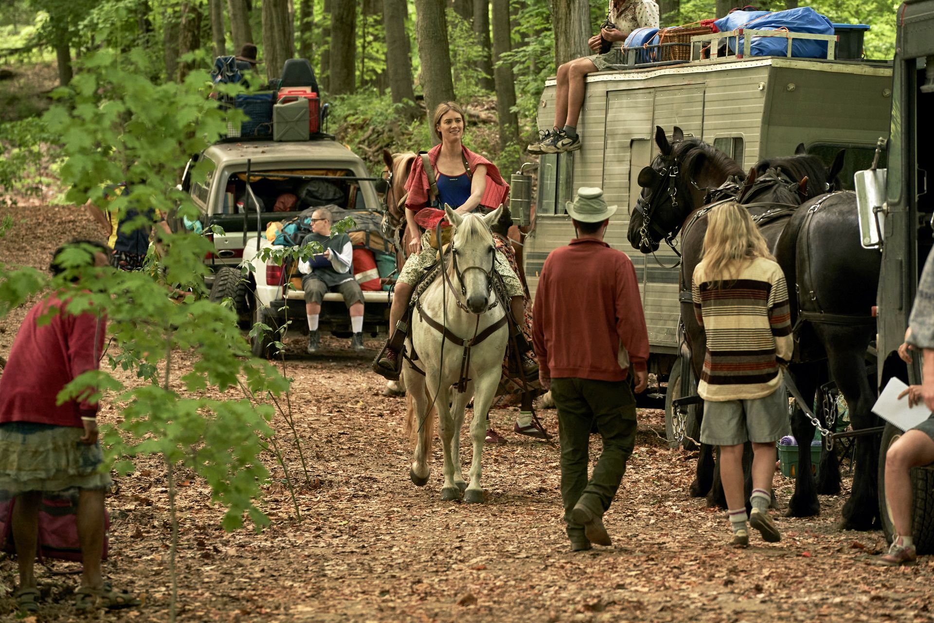 Kirsten (Mackenzie Davis) rides a horse in the woods. There are people walking around her, and in the background there are cars and campers loaded up with bags. 