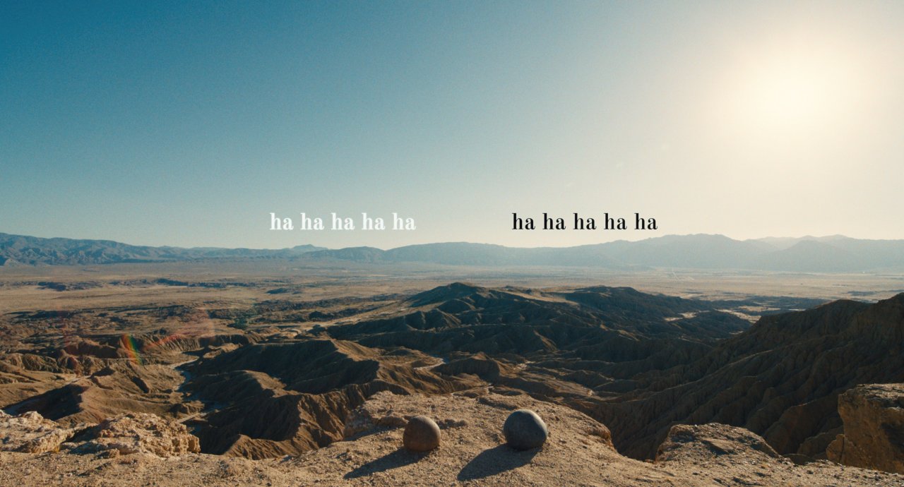 Two, large rocks sit in the foreground of a mountainous landscape. At the horizon line, above each rock, there is text that reads, "ha ha ha ha ha," as if the rocks were laughing.